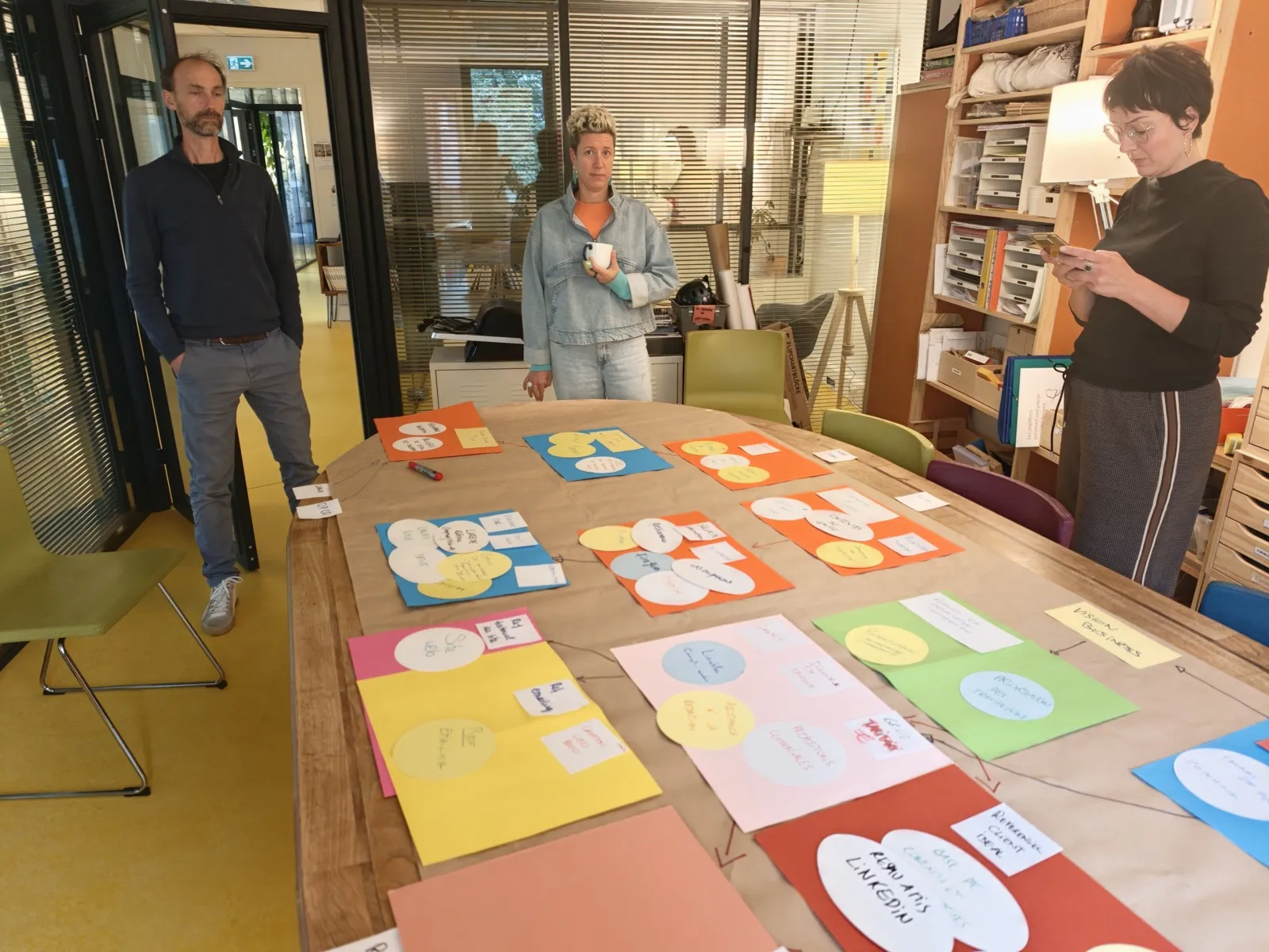 Trois personnes debout autour d'une grande table couverte de papier kraft, sur laquelle sont posés des rectangles de couleur portant des carrés et des ronds de papier annotés, reliés par des flèches tracées au feutre.